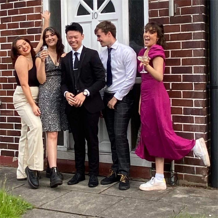 five students all dressed up some with drinks looking very happy outside the front door of their shared house
